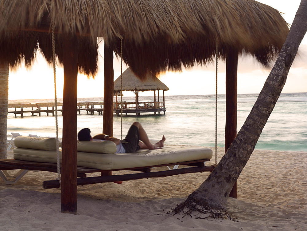 Man lying on swinging daybed under palapa watching sunrise, Mayan Riviera, Akumal, Yucatan, Quintana Roo, Mexico, North America