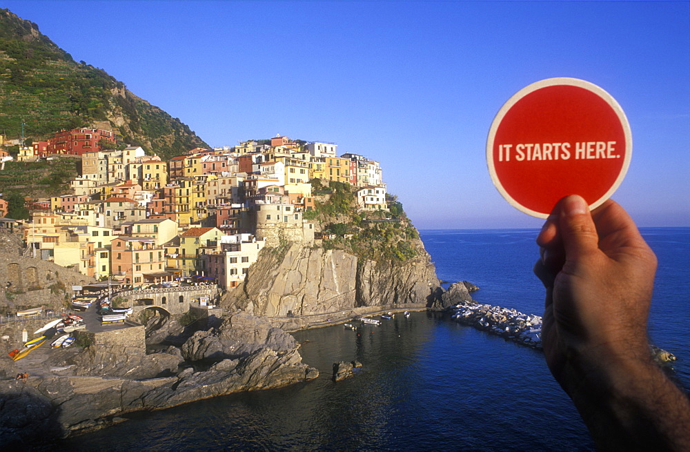 It starts here sign in front of fishing village with multi colored houses facing the sea, Manarola, Cinque Terre, UNESCO World Heritage Site, Liguria, Italy, Europe