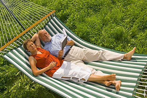 Senior citizen couple lying in a hammock, woman listening to music, man reading a book