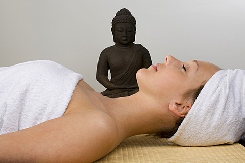 Young woman next to a Buddha statue, towel wrapped around her head, relaxing on a bamboo mat