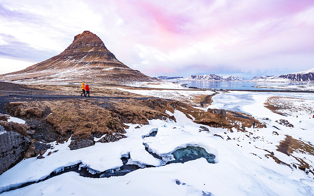 Hikers, Kirkjufell mountain, Kirkjufellfoss waterfall frozen up in front, cloudy sky with sunset, Grundarfjordur fjord, western Iceland, Iceland, Europe