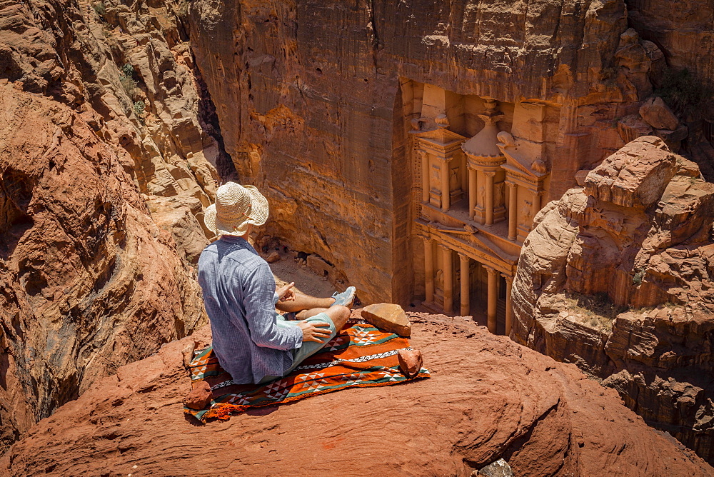 Tourist with sun hat sits on carpet and looks from above into the gorge Siq, Pharaoh's treasure house carved into rock, facade of the treasure house Al-Khazneh, Khazne Faraun, mausoleum in the Nabataean city of Petra, near Wadi Musa, Jordan, Asia