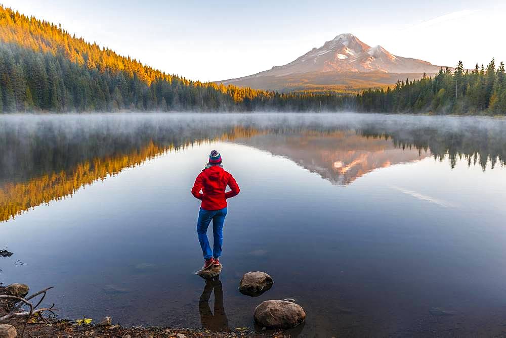 Young woman looking at view of volcano Mt. Hood with reflection in lake Trillium Lake, at sunrise, Oregon, USA, North America