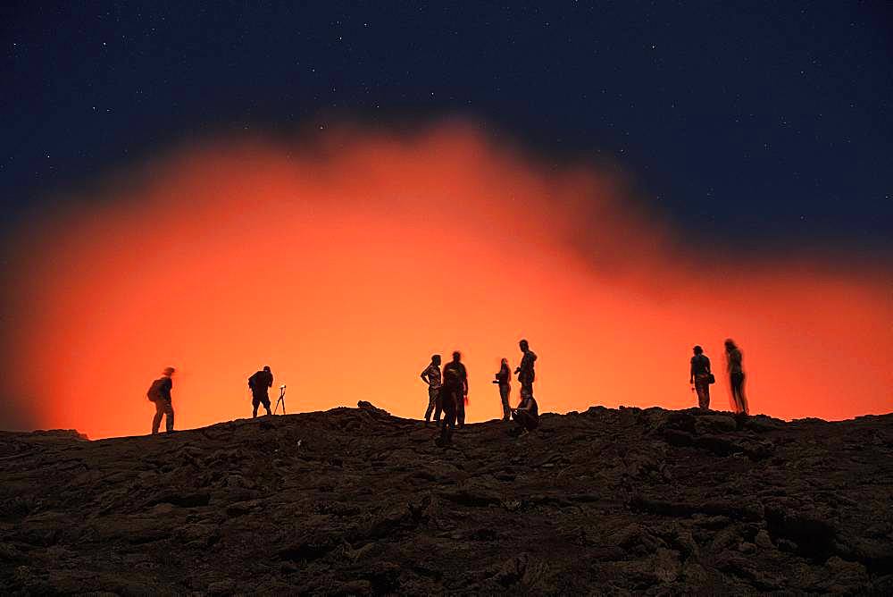 Tourists at the crater rim of the volcano Erta Ale in front of a red glowing steam cloud, Danakil Desert, Ethiopia, Africa