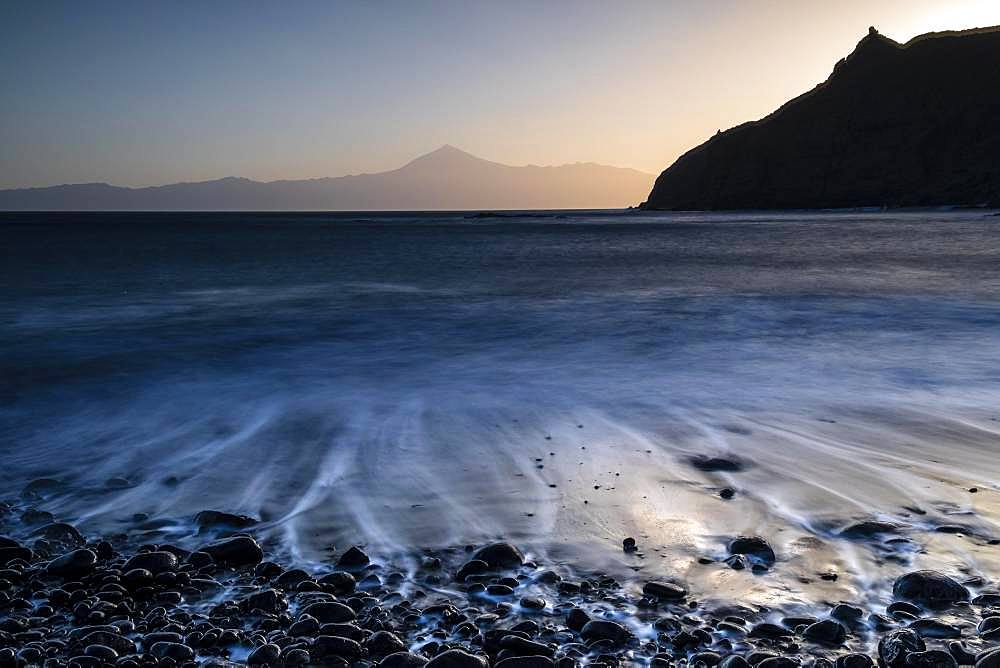 Sunrise at the beach Playa de Caleta, stones in the water, waves running out, view to Teide on Tenerife, La Gomera, Canary Islands, Spain, Europe