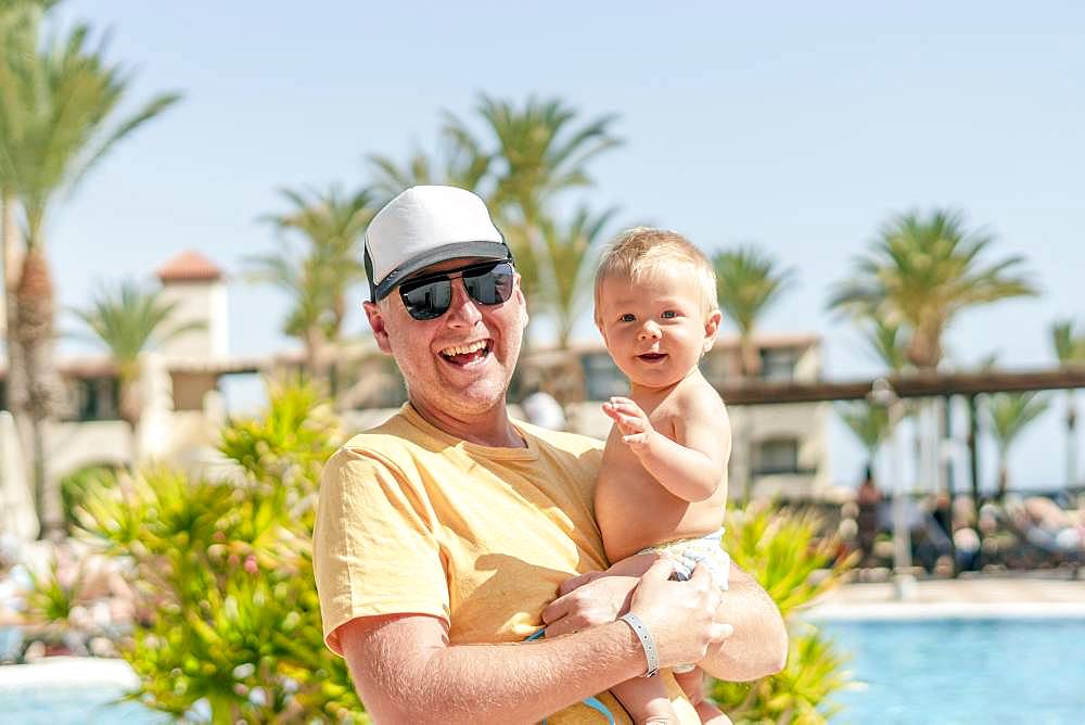 Happy father holding his cheerful son on holiday, resort with pool and palm trees as background, Portugal, Europe