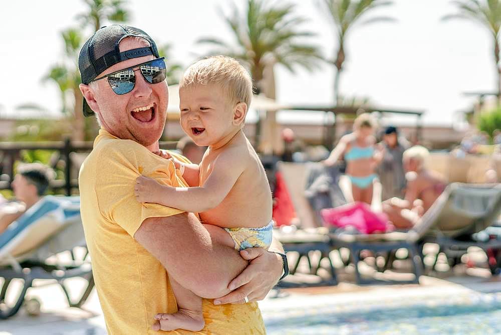 Happy father holding his cheerful son on holiday, resort with pool and palm trees as background, Portugal, Europe