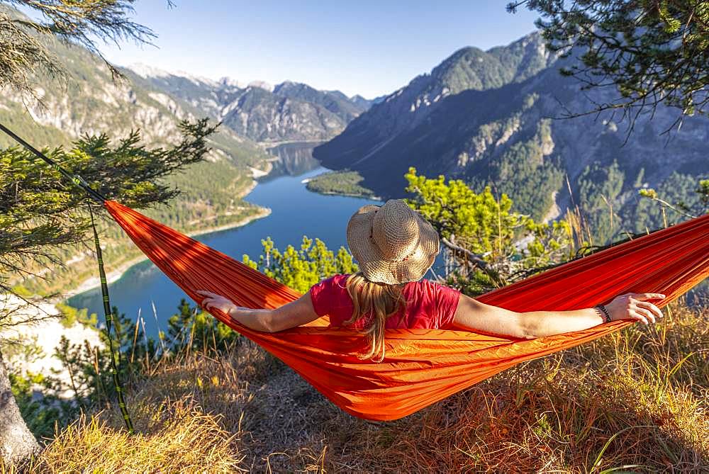 Woman with a sun hat sitting in an orange hammock, panoramic view of mountains with lake, Plansee, Tyrol, Austria, Europe