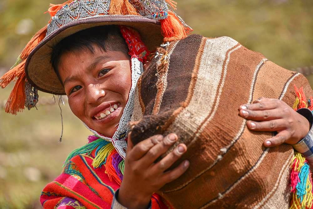 Indio boy with hat with colorful ribbons and poncho wearing sack with potatoes, portrait, at Cusco, Peru, South America