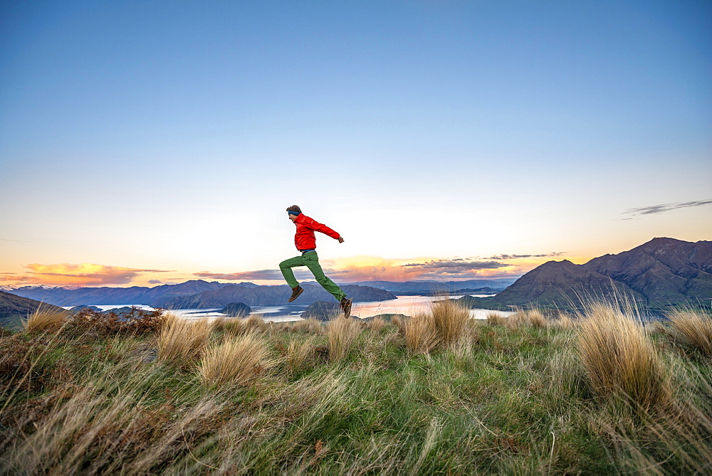 Hiker jumps into the air, panoramic view of Wanaka Lake and mountains at sunset, Rocky Peak, Glendhu Bay, Otago, South Island