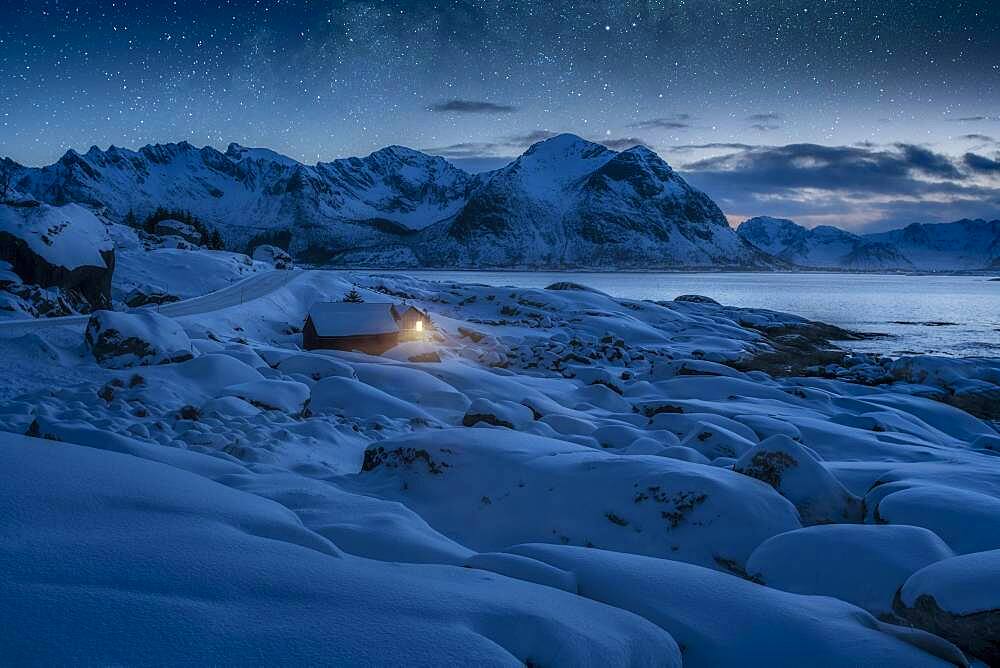 Deep snowy winter landscape by the sea, wooden cabin with warm light on the coast, behind mountains under starry sky, Kleppstad, Nordland, Lofoten, Norway, Europe