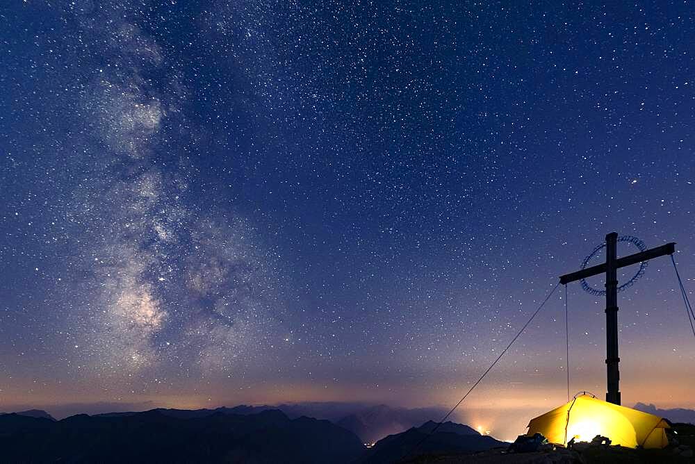 Night sky with stars and milky way over Lechtal mountains, in the foreground summit cross of the Geierkopf with tent, Reutte, Ammergau Alps, Tyrol, Austria, Europe