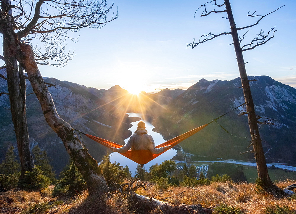 Young man sitting in a red hammock, panoramic view of mountains with lake, sun star, sunset, Plansee, Tyrol, Austria, Europe