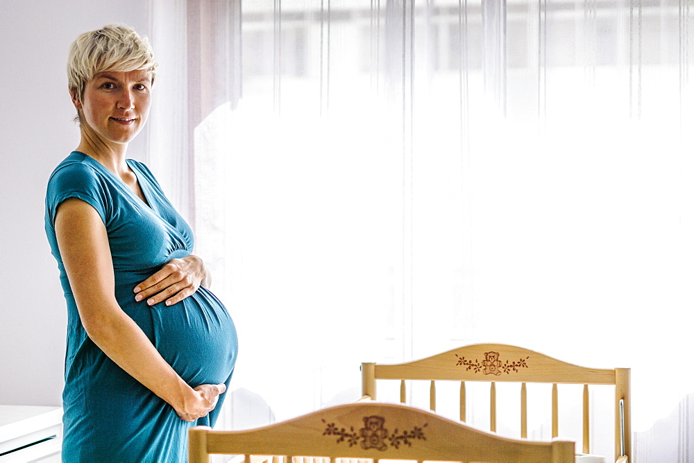 Pregnant woman holding her belly by baby cot next to the window