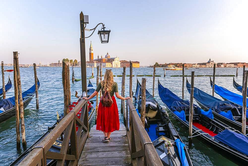 Young woman with red dress on a jetty, Venetian gondolas, in the back church San Giorgio Maggiore, Venice, Veneto, Italy, Europe