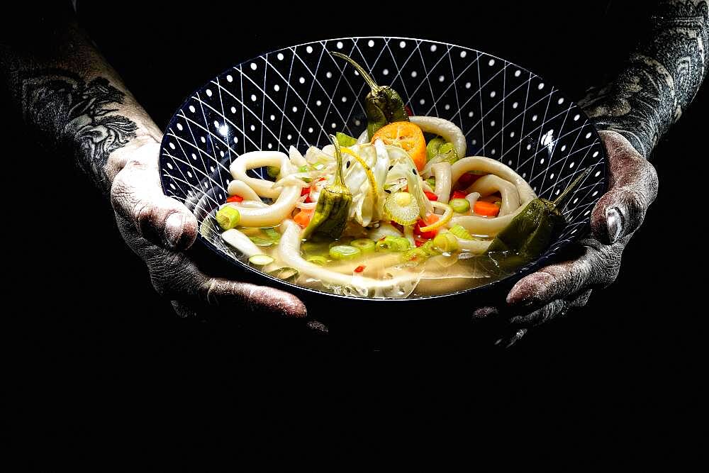 Tattooed hands hold plate with udon bowl, japanese noodle soup