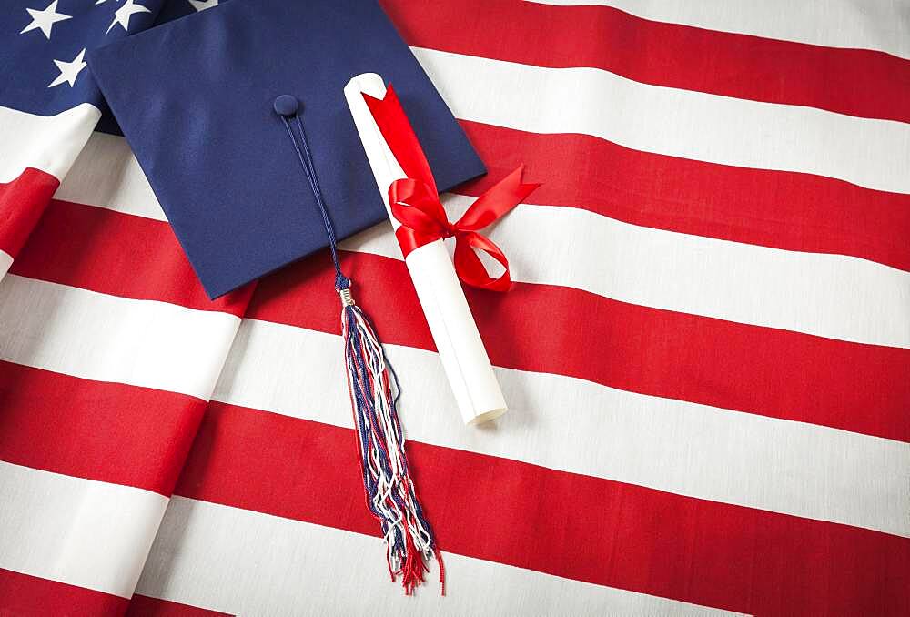 Graduation cap with tassel and red ribbon wrapped diploma resting on american flag