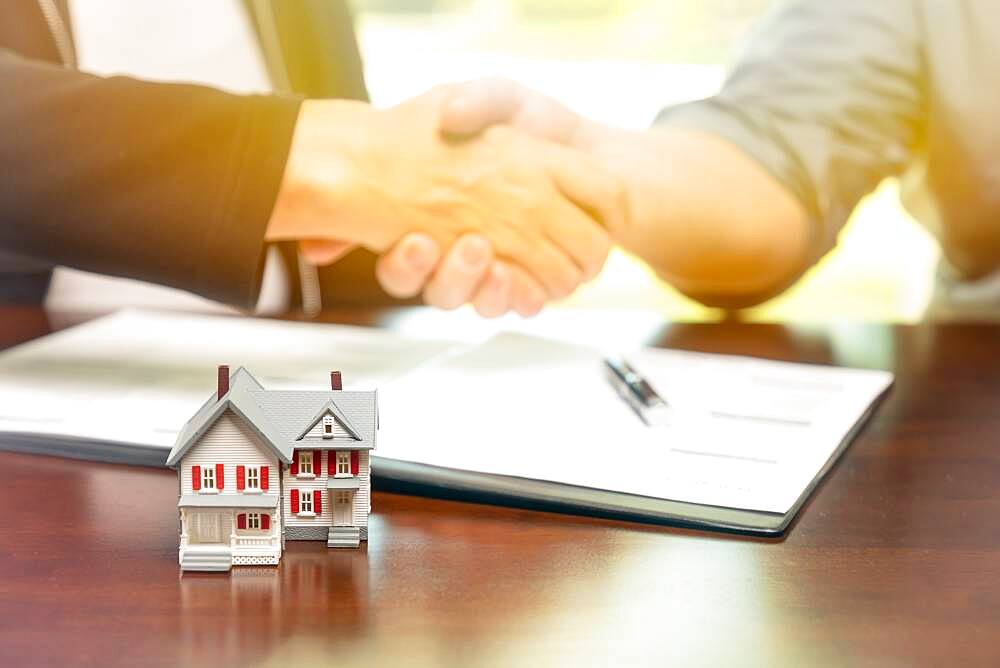 Real estate agent and customer sign contract papers and shake hands with small model home in front