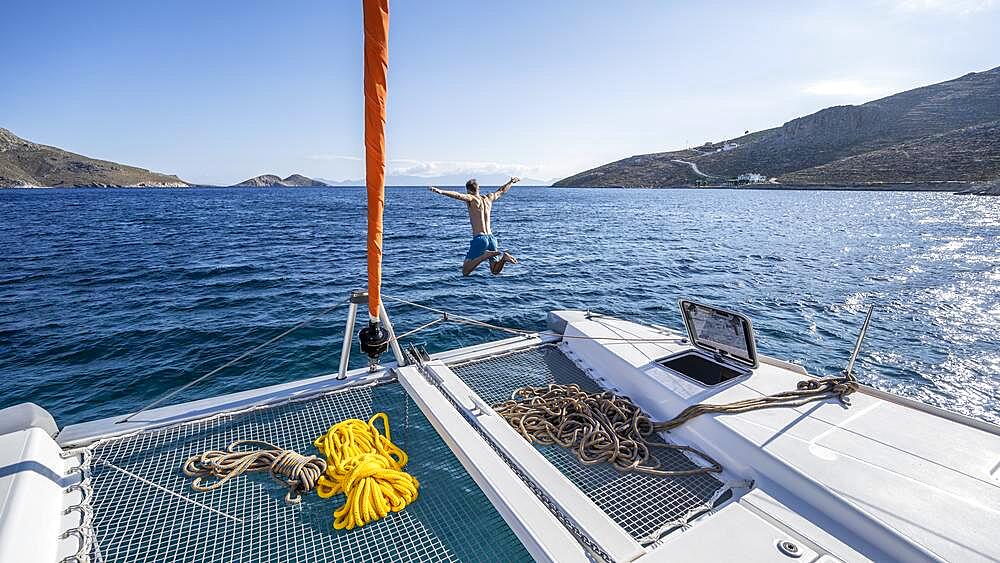 Young man jumps into the water, ropes on a sailing catamaran, sailing trip, Tilos, Dodecanese, Greece, Europe