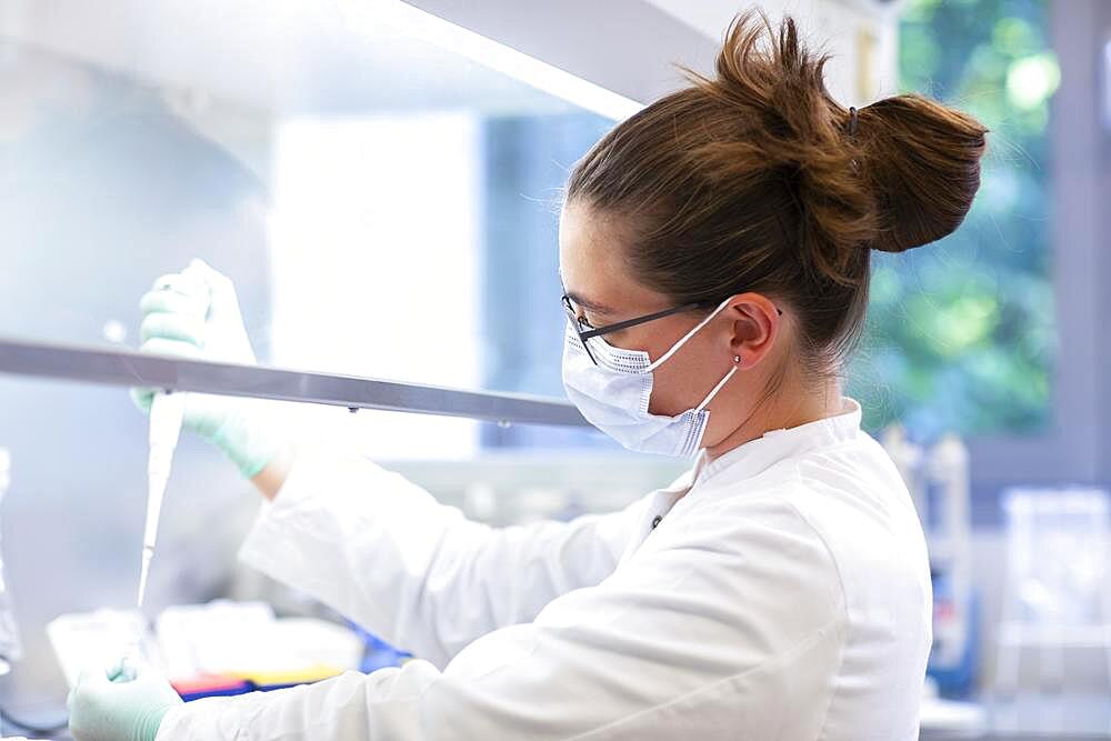 Young lab technician wearing a face mask and pipetting a sample in a laboratory with laboratory equipment at a workflow, Freiburg, Baden-Wuerttemberg, Germany, Europe