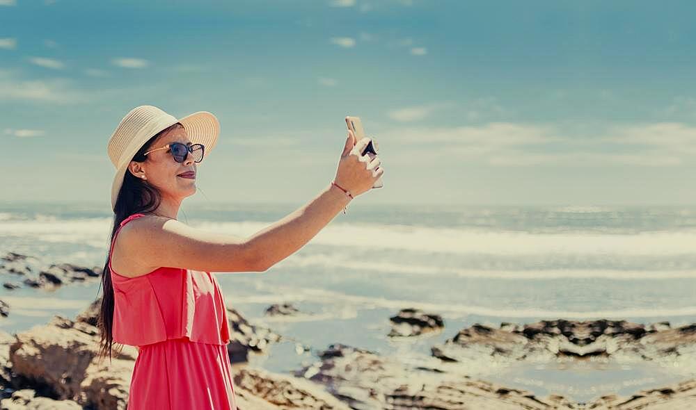 Hat woman taking photos at sea, latin girl taking vacation photos, rear view of girl taking selfie on the beach