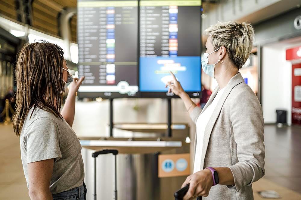 Two travelling women wearing protective masks discussing by flight information board at the Faro airport