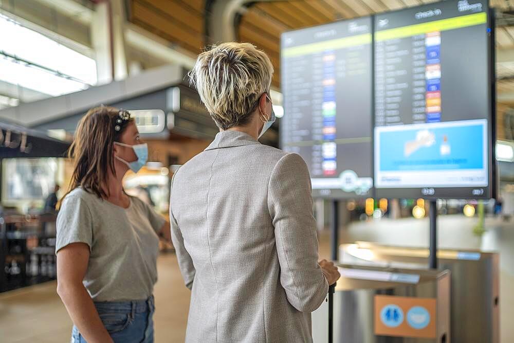 Two travelling women wearing protective masks discussing by flight information board at the Faro airport