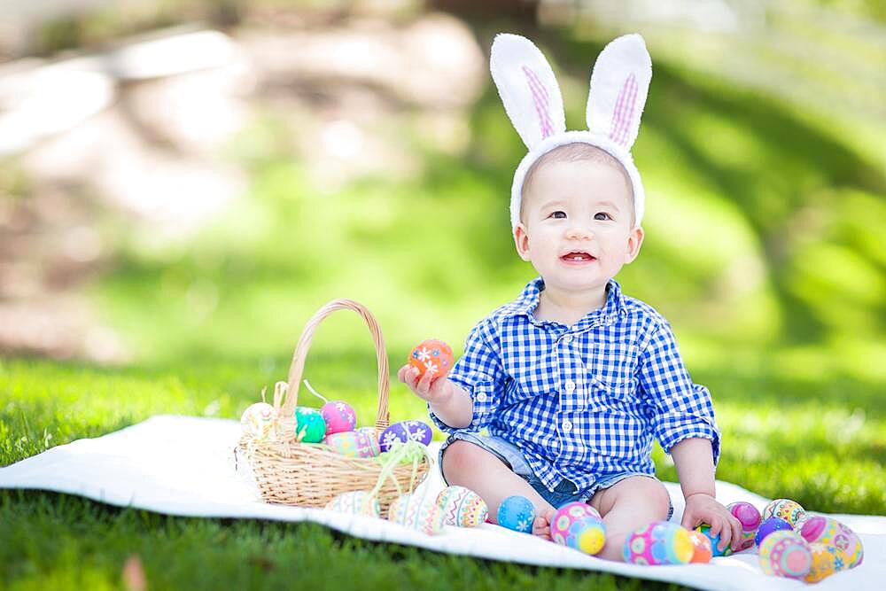 mixed-race chinese and caucasian baby boy outside wearing rabbit ears playing with easter eggs