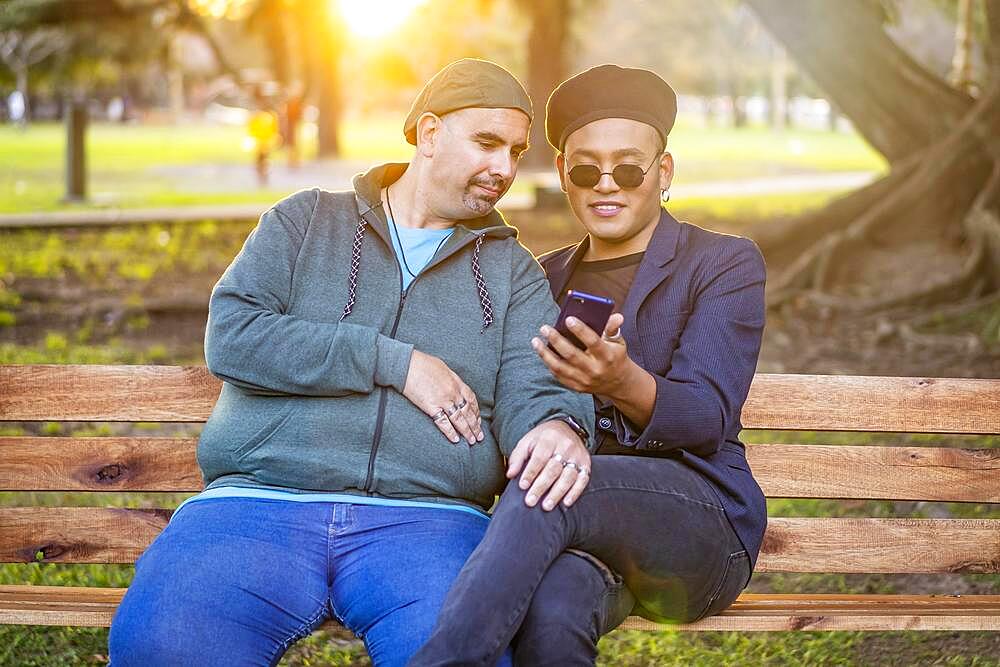 Gay Latino male couple sitting on a bench in a park at sunset, wearing fashionable hats, holding a cell phone, smiling