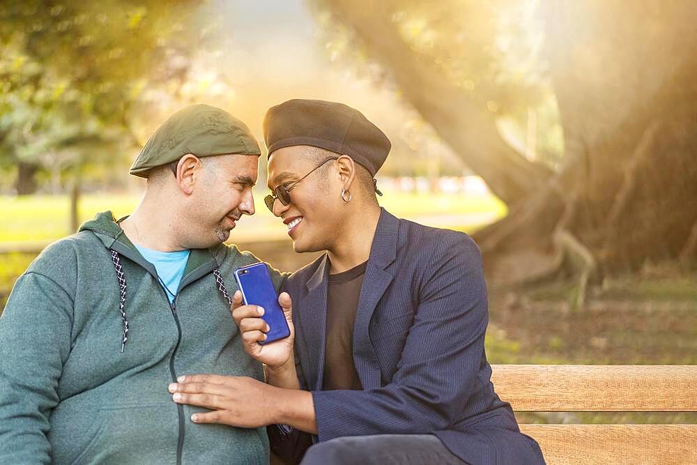 Gay Latino male couple sitting on a bench in a park at sunset, wearing fashionable hats, holding a cell phone, smiling