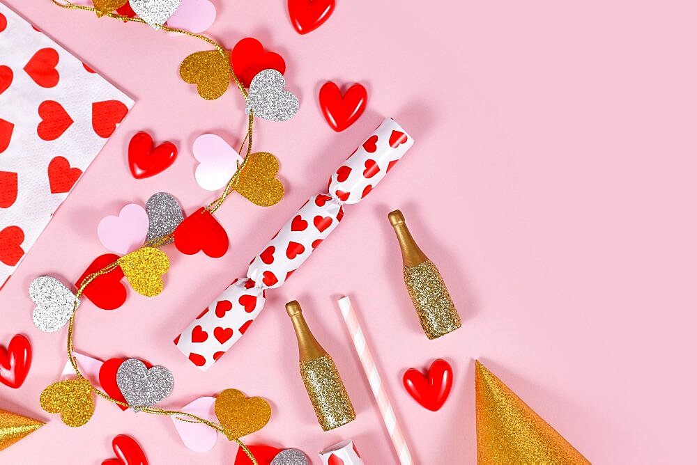 Party flat lay with heart garland, napkin, champagne bottles and party hats on pink background