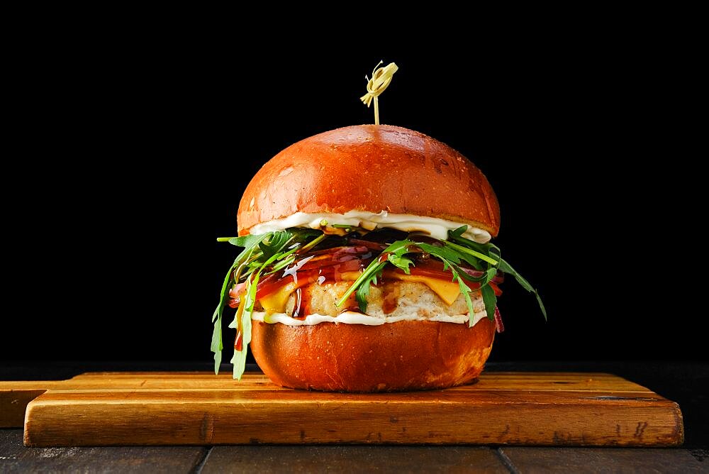 Burger with chicken patty teriyaki sauce on wooden serving board over black background