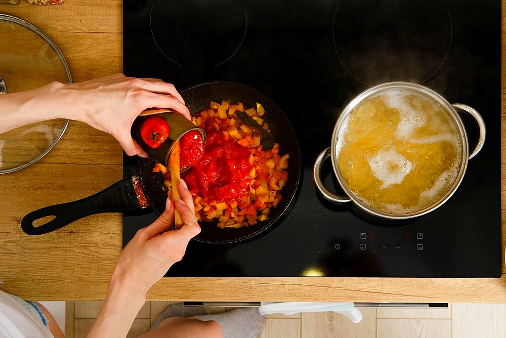 Closeup overhead view of female hand adding canned whole peeled tomatoes into frying pan while preparing pasta sauce