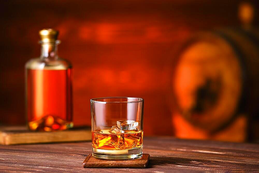 Glass of whiskey with ice on wooden table with bottle and barrel on background