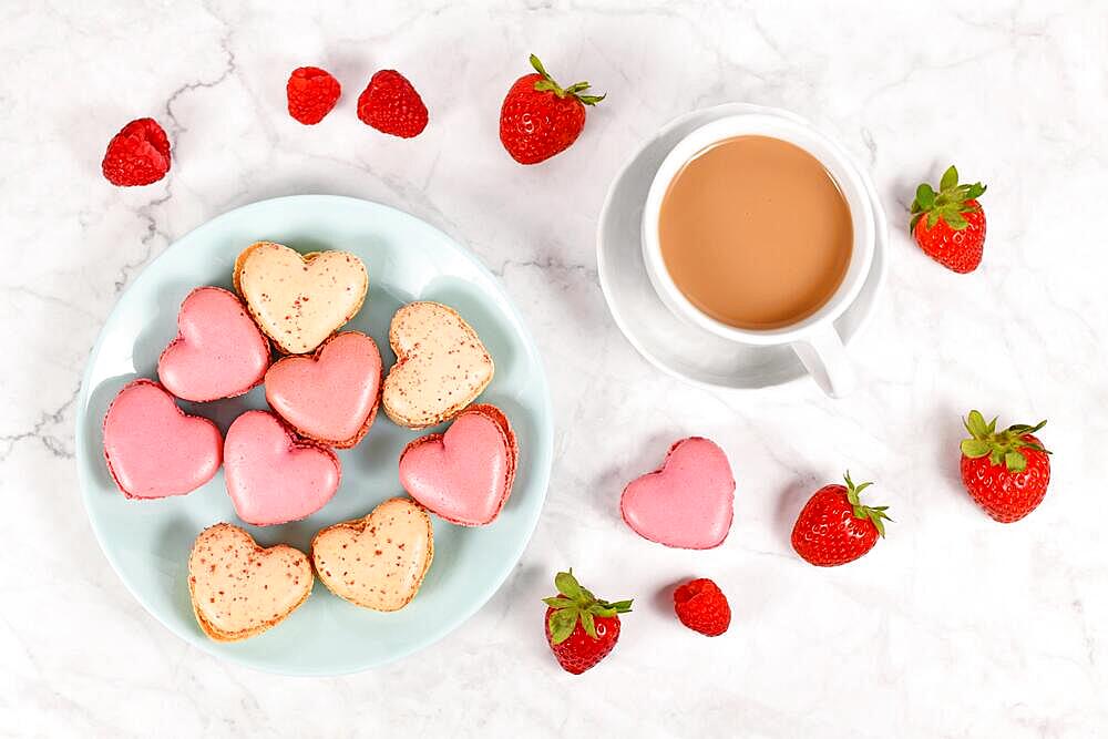 Pink and beige heart shaped French macaron sweets next to coffee cup and strawberry and raspberry fruits