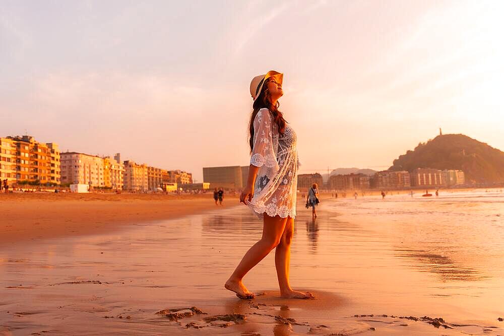 A woman in the sunset in a white dress with a hat walking by the sea