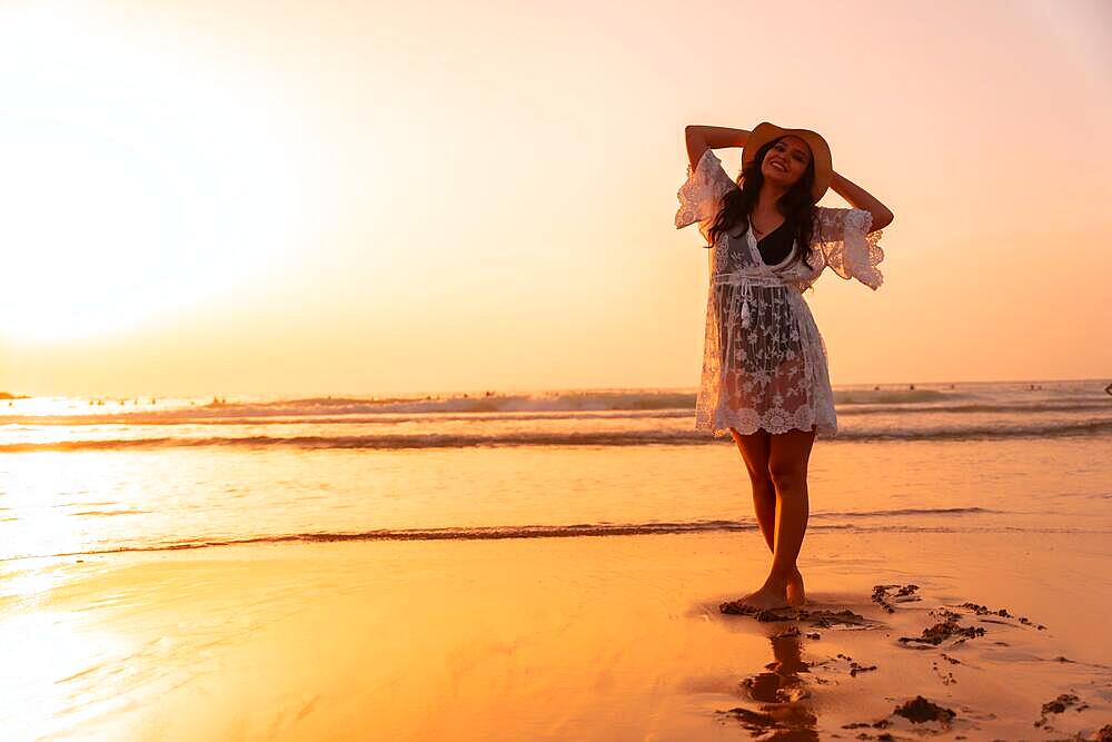 A woman in the sunset in a white dress at the sea in summer with a hat