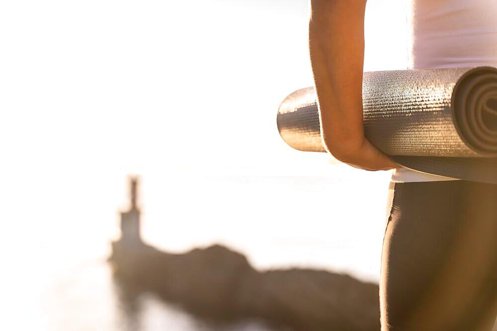 Hands of a woman with the yoga mat in nature by the sea at sunset, healthy and naturist life, pilates outdoors