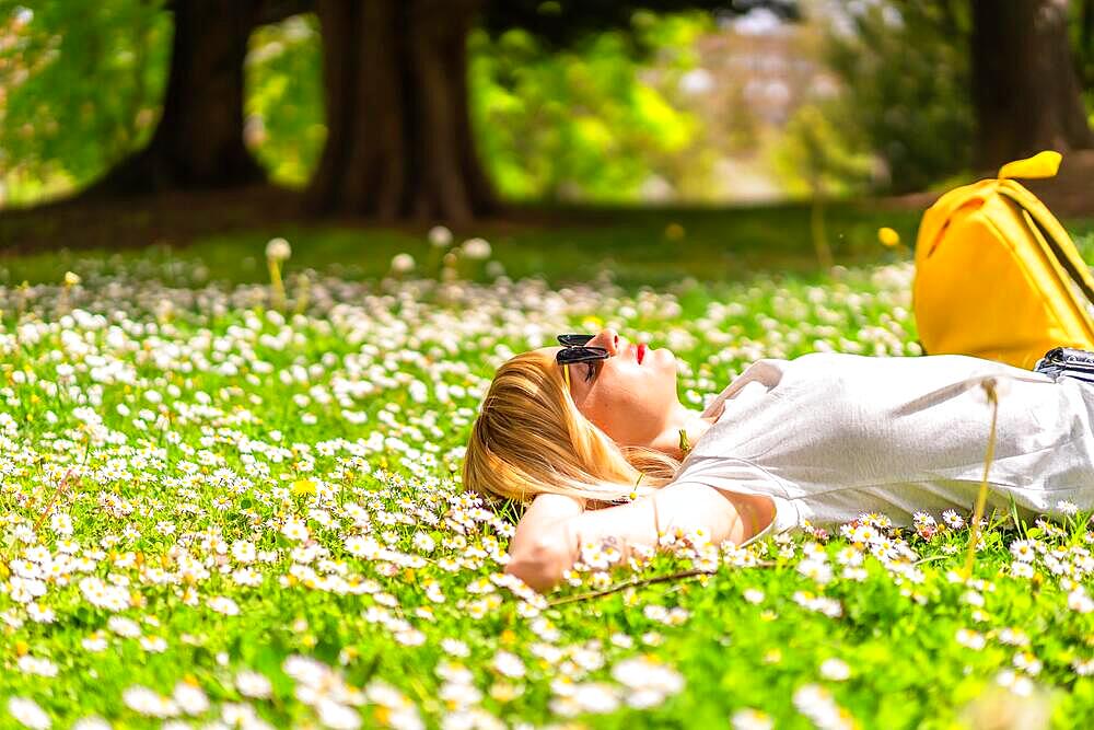 A young blonde girl in a hat unwinding and breathing pure hair in the spring in a park in the city, nature, lying on the grass next to daisies, looking at the camera