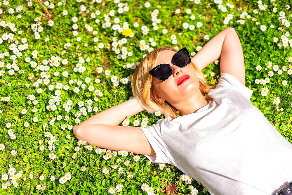 A young blonde girl in a hat and sunglasses breathing pure haire in the spring in a park in the city, nature, lying on the grass next to daisies, part copy space