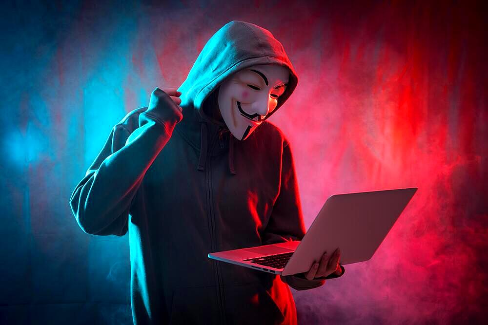 Hacker with an anonymous mask with a computer and making a fight symbol, with a background of smoke and colored leds