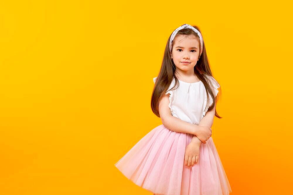 Cute little girl stands in a yellow-tinted studio, looking into the camera with innocence. Her long hair and fashionably adorned dress reflect her childhood as she captures this moment of beauty