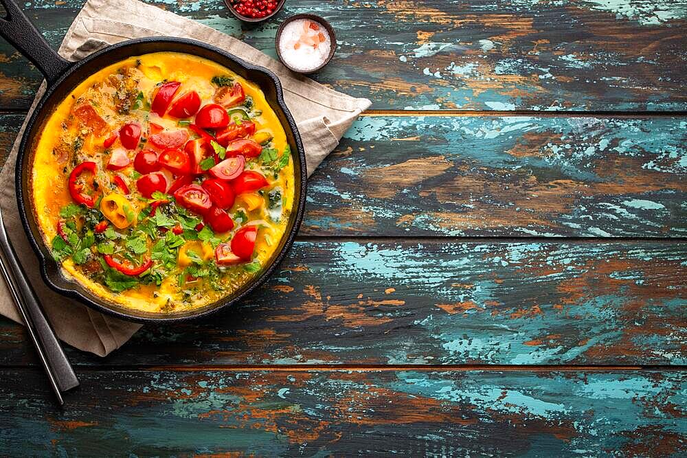 Healthy frittata in cast iron pan with fried beaten eggs and seasonal vegetables on rustic wooden background. Italian omelette with organic spinach, bell pepper, tomatoes, top view, copy space