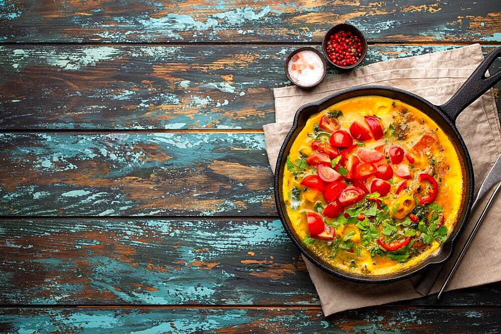 Healthy frittata in cast iron pan with fried beaten eggs and seasonal vegetables on rustic wooden background. Italian omelette with organic spinach, bell pepper, tomatoes, top view, copy space