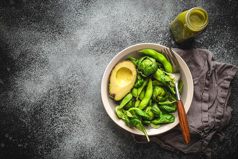 Green healthy salad with spinach, brussels sprouts, avocado in bowl, green detox smoothie in a bottle on gray stone background, top view. Raw food diet and clean eating, vegan healthy meal, copy space