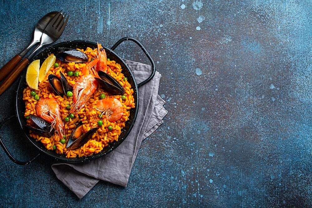 Classic dish of Spain, seafood paella in traditional pan on blue stone background top view. Spanish paella with shrimps, clamps, mussels, green peas and fresh lemon wedges from above, space for text