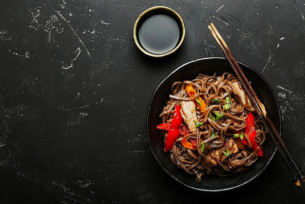 Bowl with Asian chicken stir fry soba noodles with vegetables on dark black stone background from above, Chinese Thai or Japanese noodles dish with soy sauce. Space for text