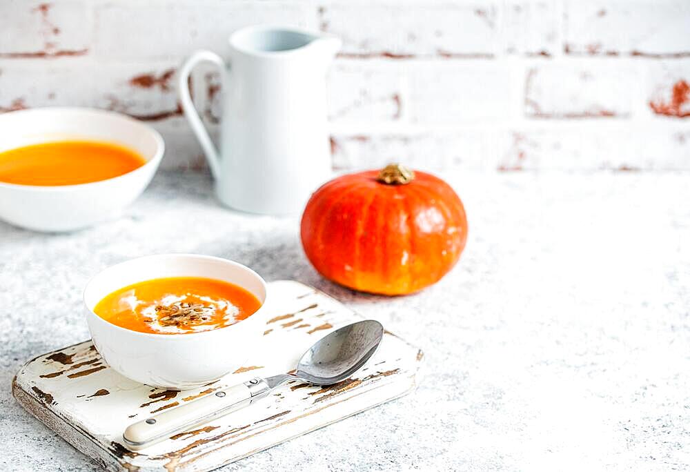 Homemade pumpkin cream soup served in white ceramic bowl on white table with spoon decorated with whole pumpkin, angle view, selective focus. Autumn cozy comfort food, background space for text