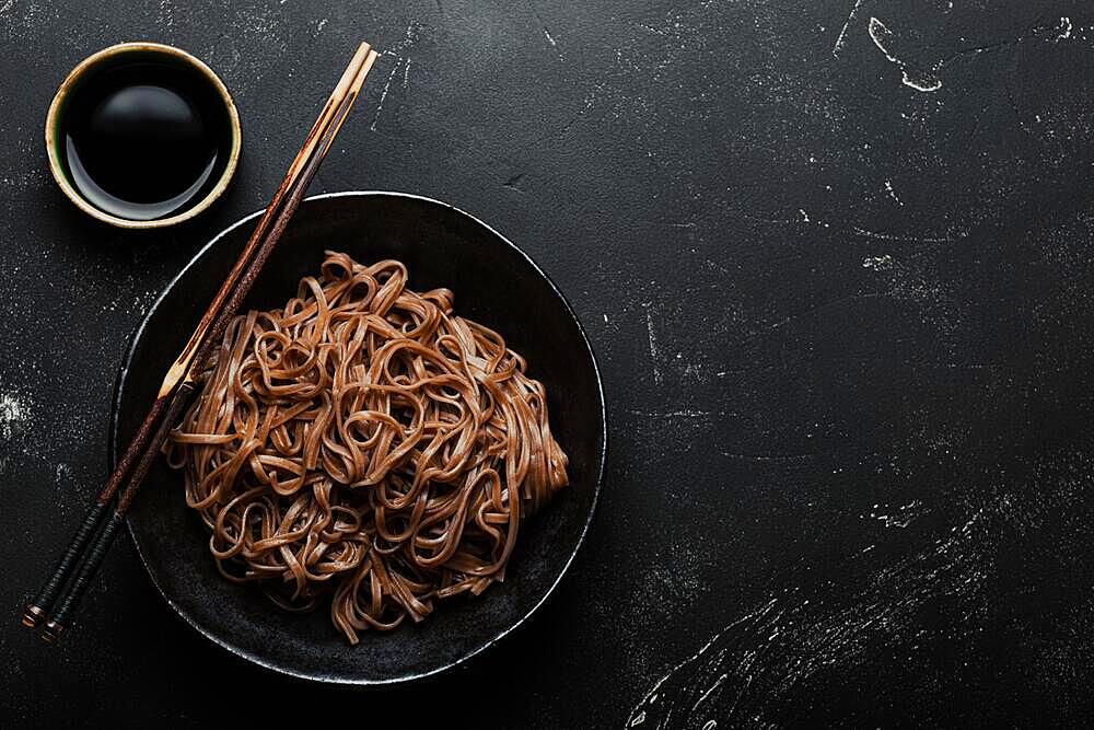 Bowl with plain soba noodles on dark black stone background from above, noodles for cooking Chinese Thai or Japanese dish with soy sauce, space for text