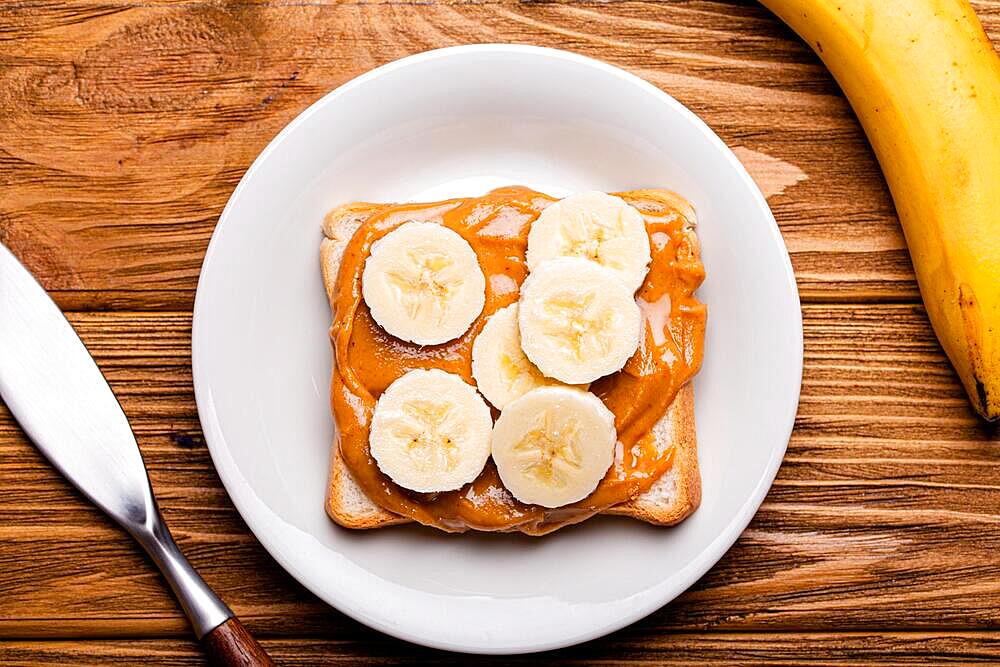 Toast with peanut butter and banana on white ceramic plate with knife on rustic wooden background, top view flat lay